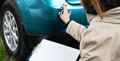 Person, die Schäden an der Autostoßstange mit einem Klemmbrett in der Hand im Freien untersucht.