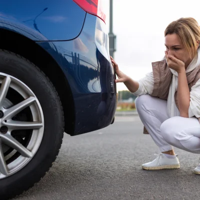Frau untersucht Schaden an der Heckstoßstange von einem blauen Auto