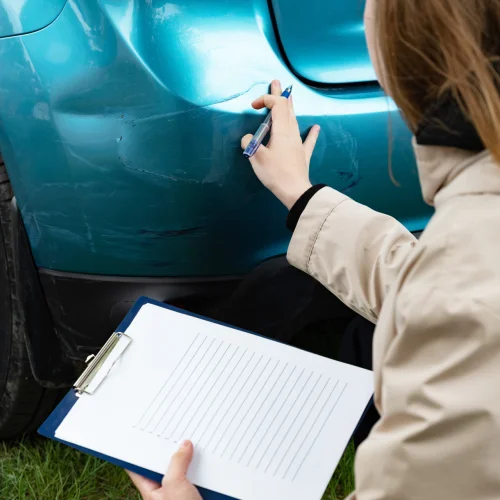 Person, die Schäden an der Autostoßstange mit einem Klemmbrett in der Hand im Freien untersucht.